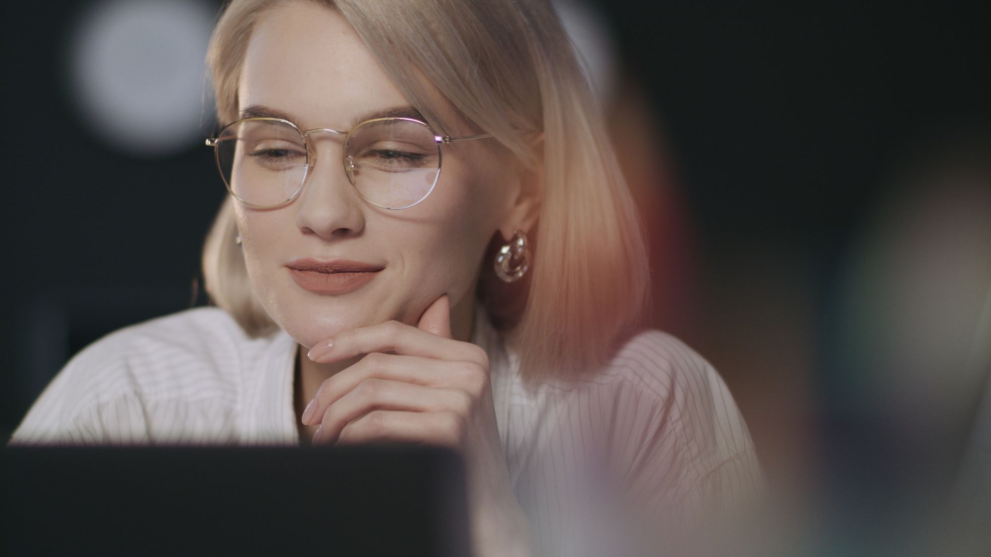 A person in glasses at a laptop in a dim office, considering the screen