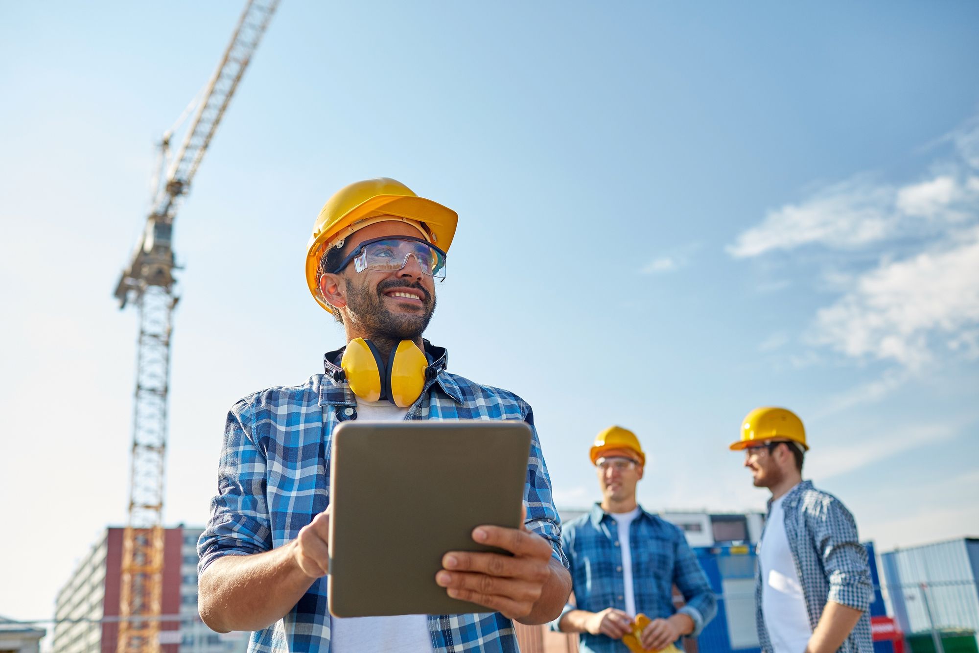 A worker with a tablet on a job site, a crane and crew behind