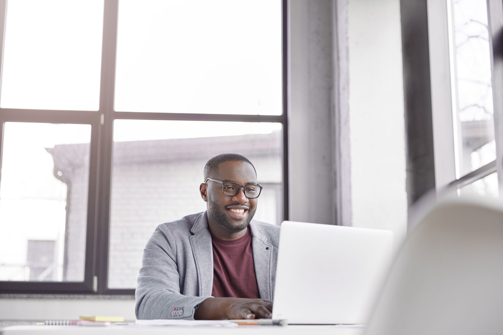 A person smiling at a laptop by a large office window
