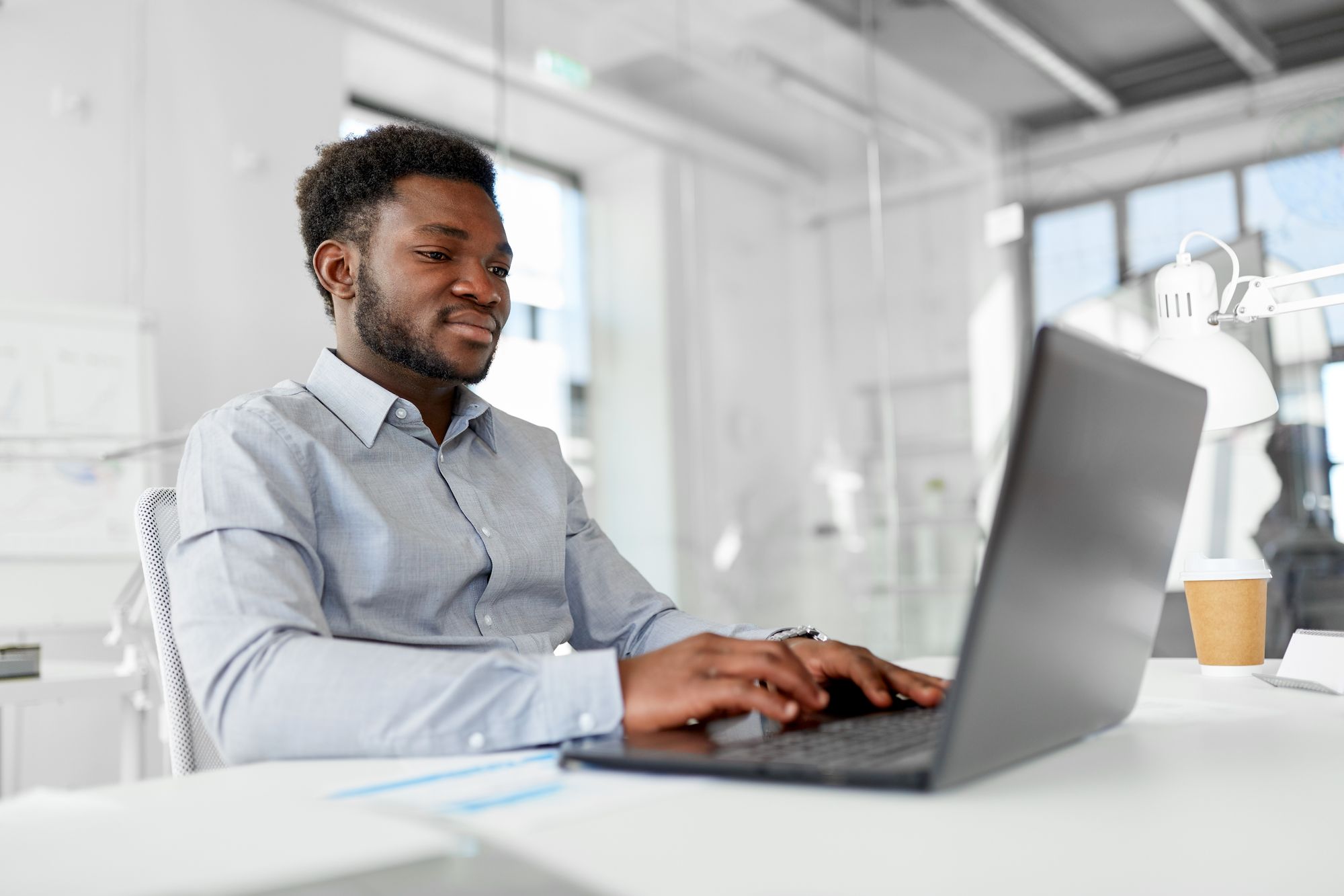A person at a laptop in a bright office, coffee beside them