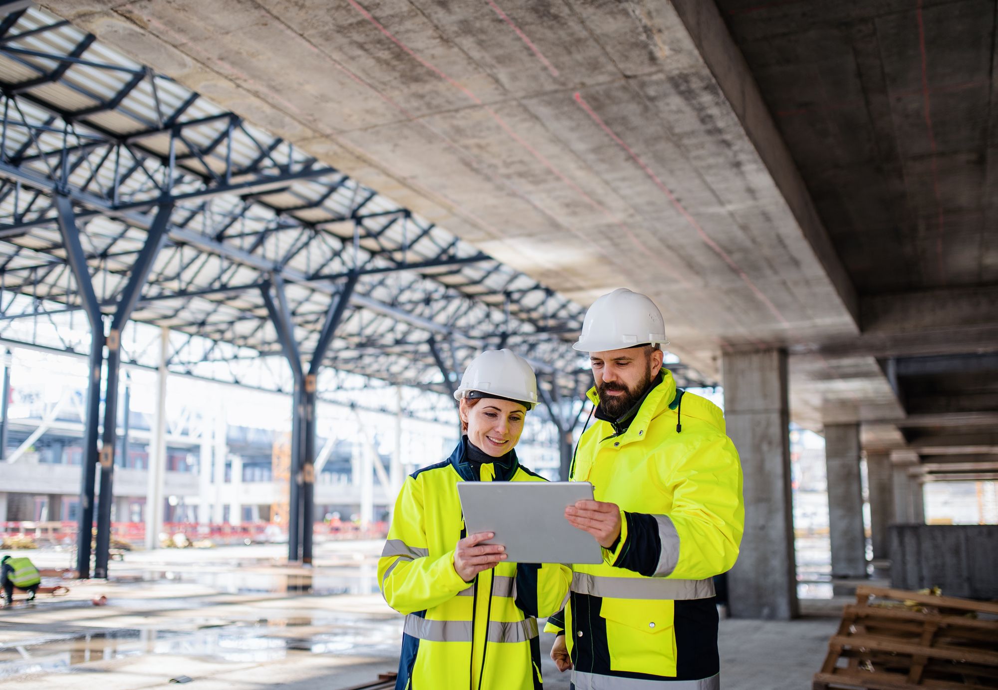 Two people in high-visibility vests looking at a tablet together under a concrete structure