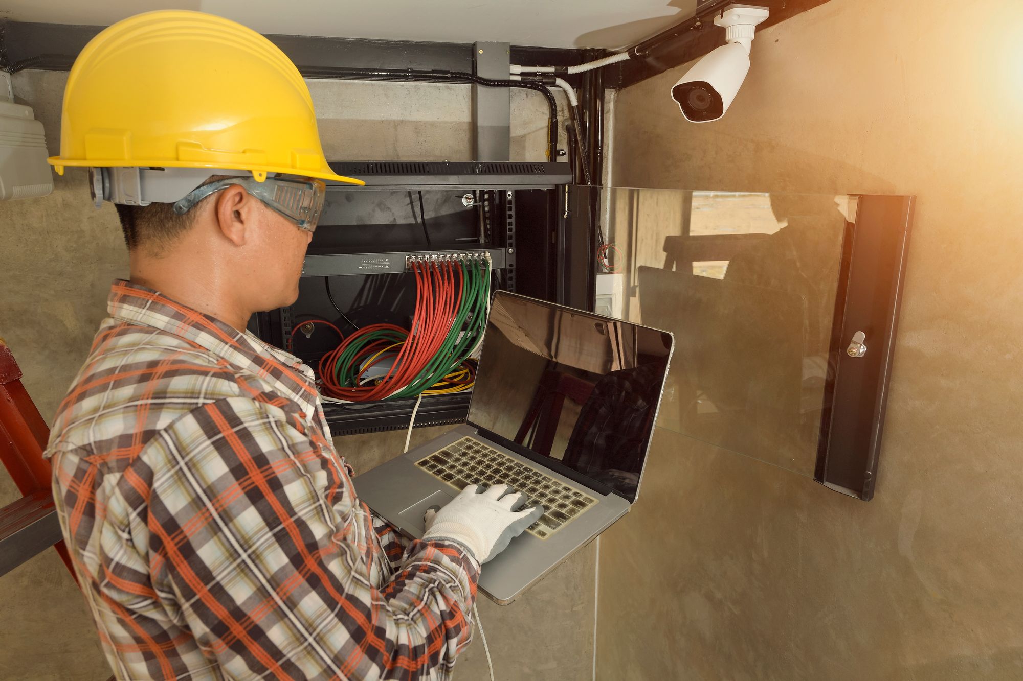 An electrician in a hard hat at a laptop in front of a network panel