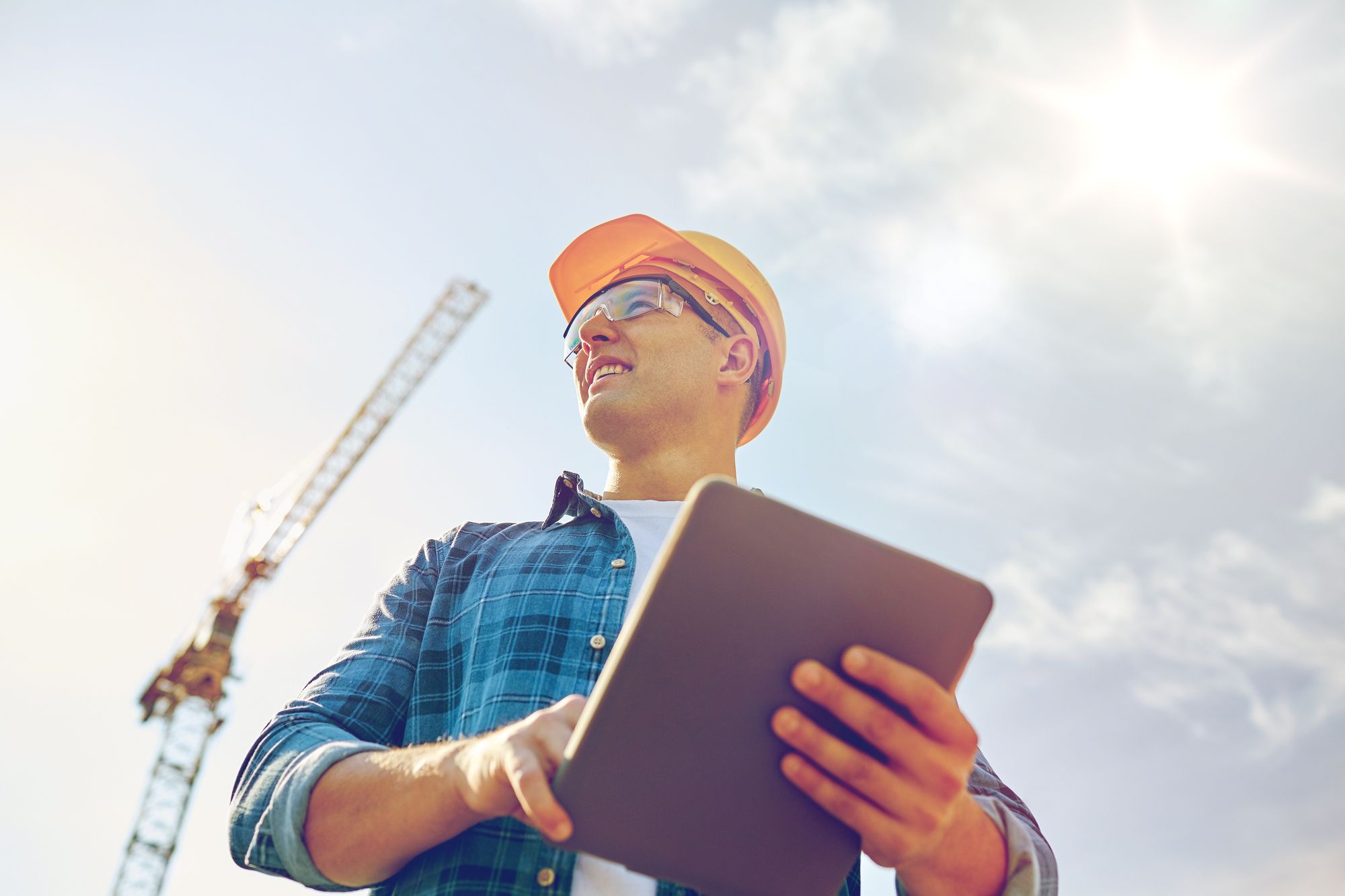A worker in a hard hat looking up from a tablet on a job site, a crane behind